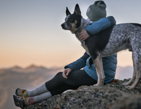 moça, com roupa de inverno, sentada no alto de uma montanha abraçada a seu cachorro