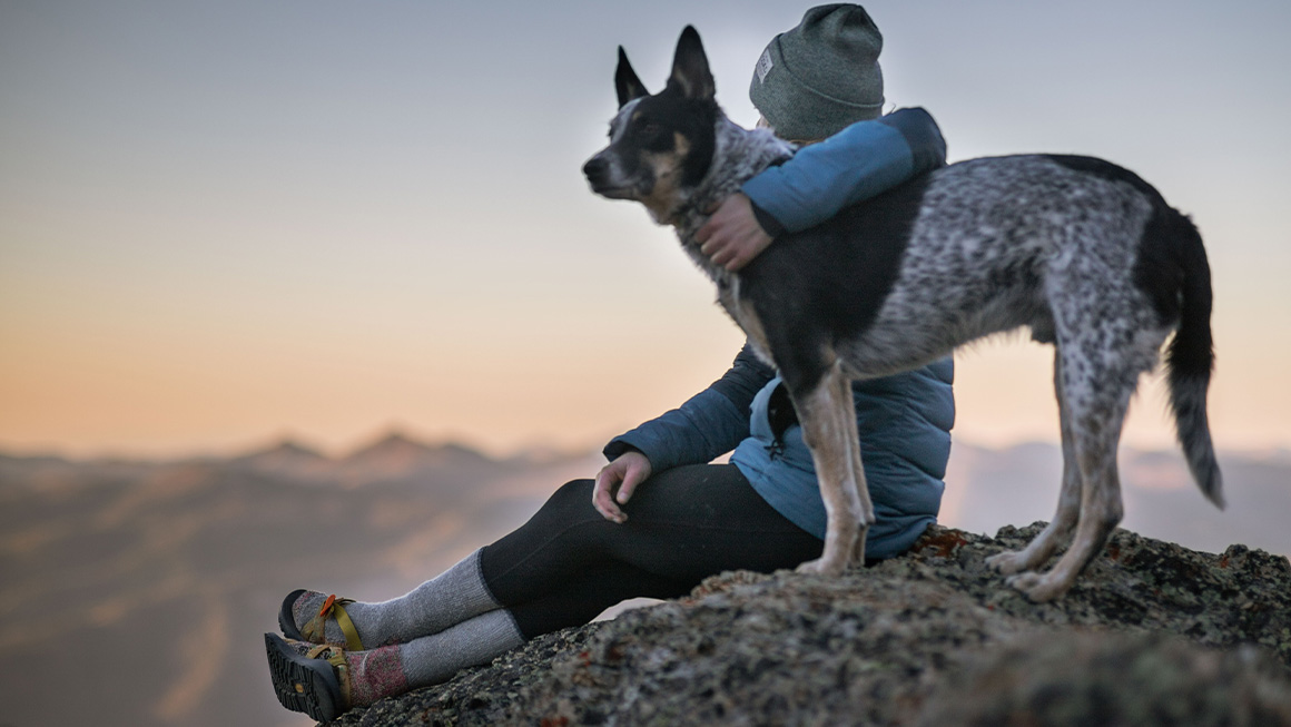 moça, com roupa de inverno, sentada no alto de uma montanha abraçada a seu cachorro