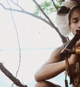 Flautista indígena sentado, tocando flauta.